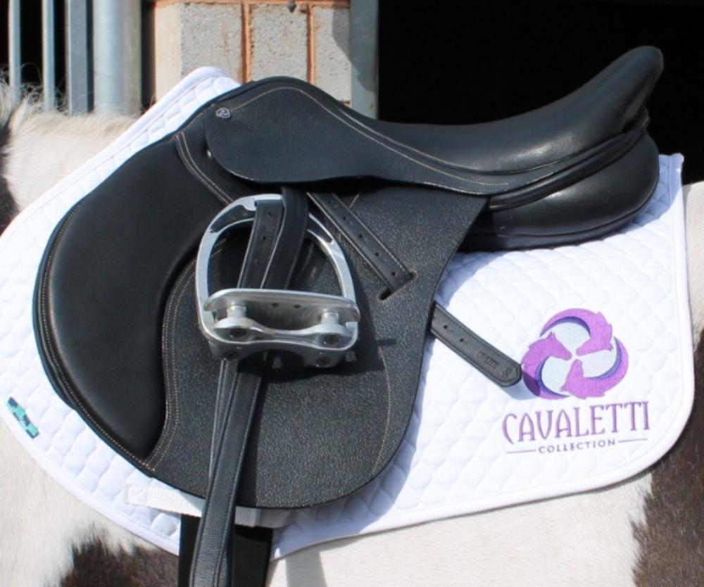 Close‑up of a black Cavaletti Collection saddle on a white quilted saddle pad on a horse with a dark‑and‑white coat.