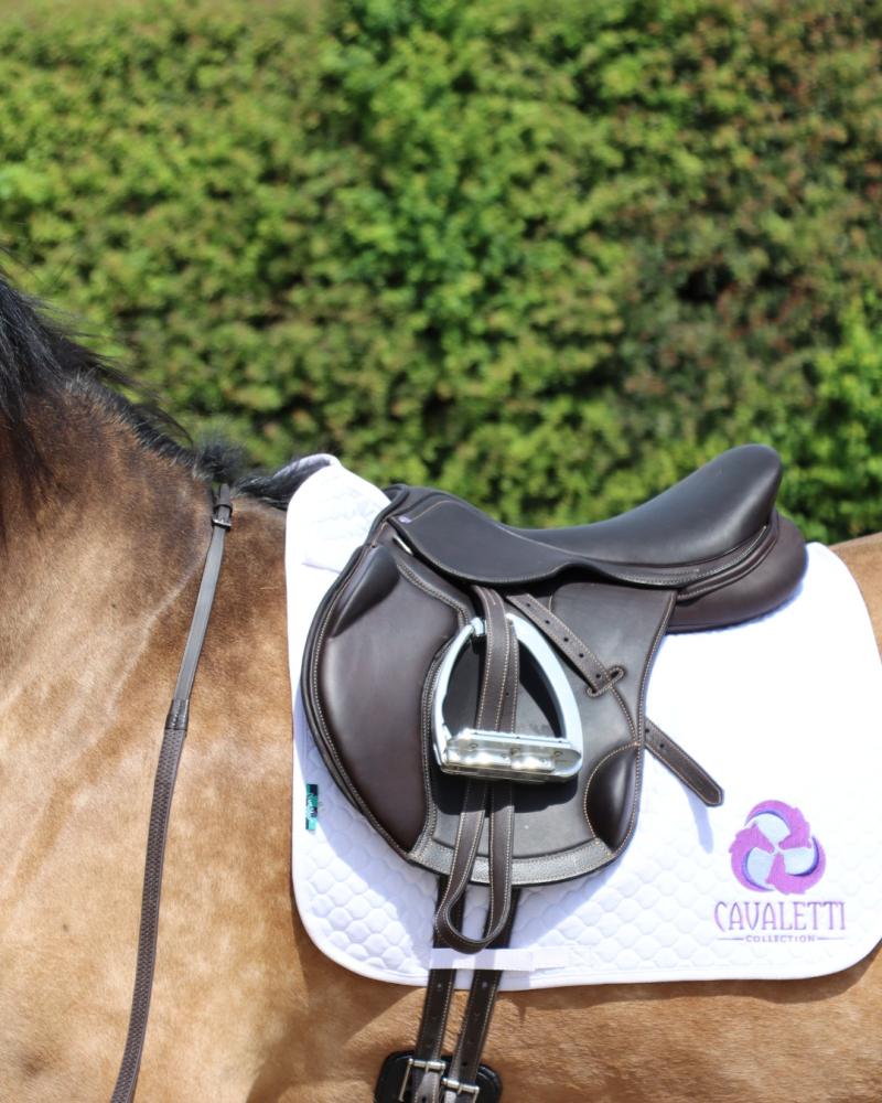 Brown Cavaletti Collection saddle on a white quilted pad in an outdoor setting with green foliage.