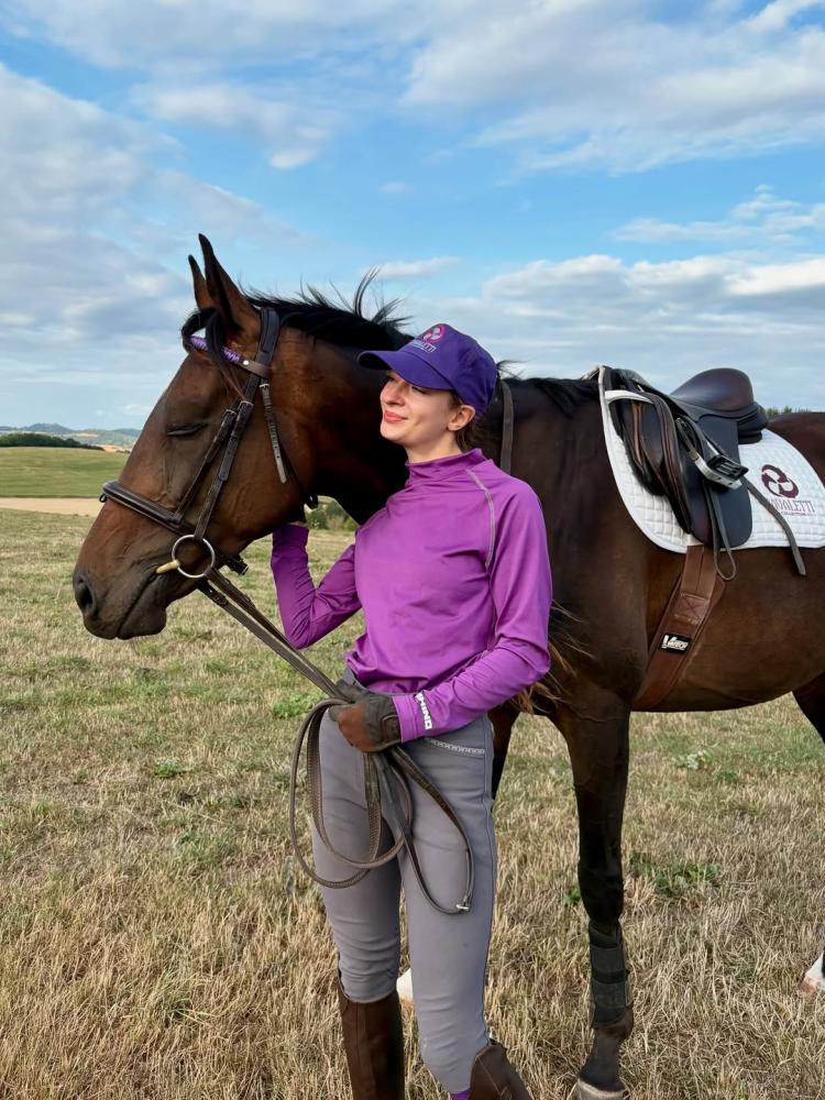 Rider standing beside a brown horse in an open grassy field, both wearing Cavaletti Collection tack. Photo credit from talesoftwomares.