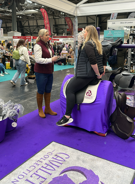 Cavaletti Collection saddle demonstration at a trade stand, with a rider testing a saddle on a purple stand during an equestrian event.