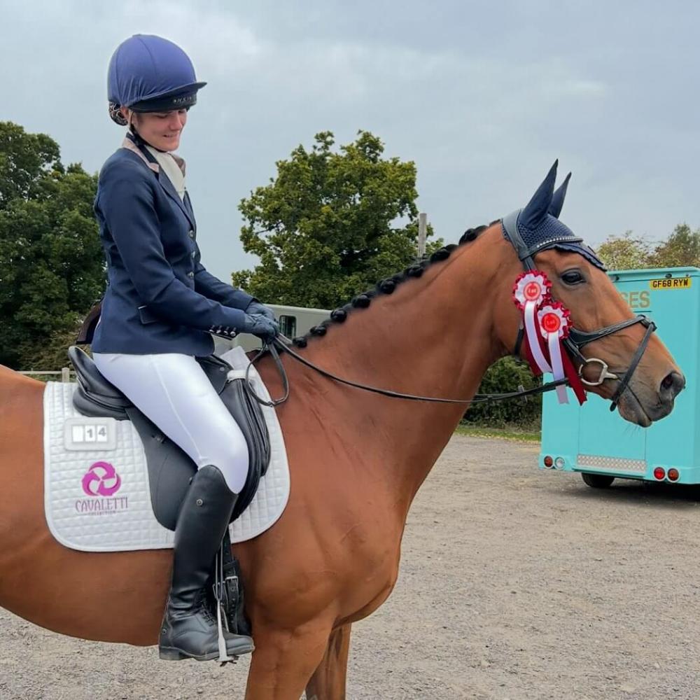 Rider in navy show jacket and white breeches sitting on a plaited bay horse wearing a navy ear bonnet red and white rosettes and a white Cavaletti saddle pad in the competition warm up area. Image credit to team-allerton