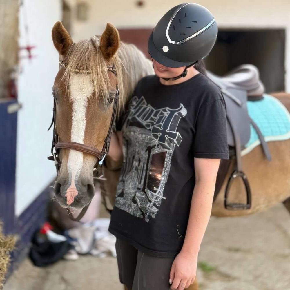 Cavaletti Collection: A rider in a black helmet stands beside a light brown horse with a white blaze, fully tacked with a turquoise saddle pad in a stable setting.