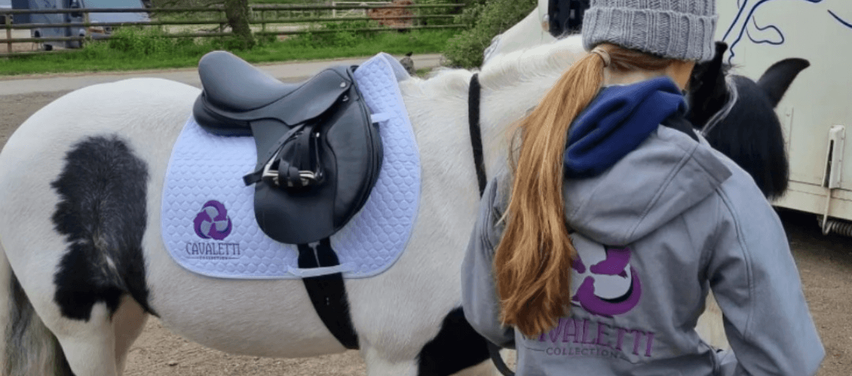 Rider standing beside a white‑and‑black horse equipped with a black saddle and white Cavaletti Collection saddle pad in an outdoor setting.
