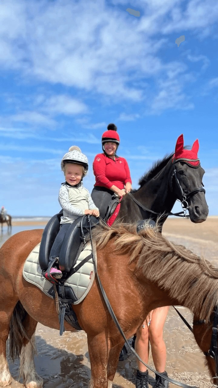 Young child riding a pony on a sunny beach beside an adult rider on a black horse both smiling and enjoying a relaxed equestrian day out. Clear blue sky and shallow water in the background. Image credit to mirandashorseyadventures